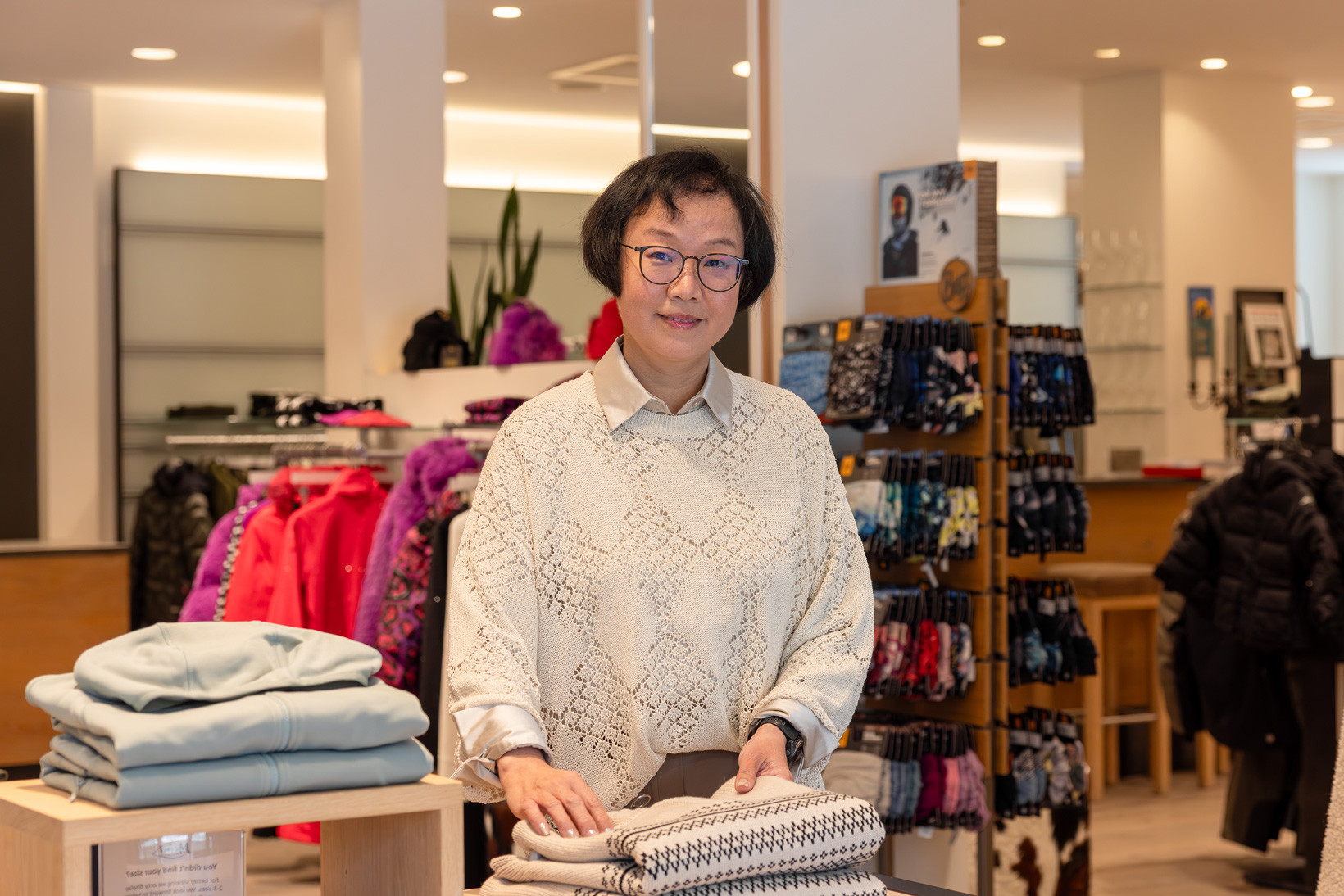 Eine lächelnde Mitarbeiterin mit Brille und hellem Strickpullover steht in einem modern eingerichteten Sport- und Modegeschäft und legt Kleidung zusammen.
