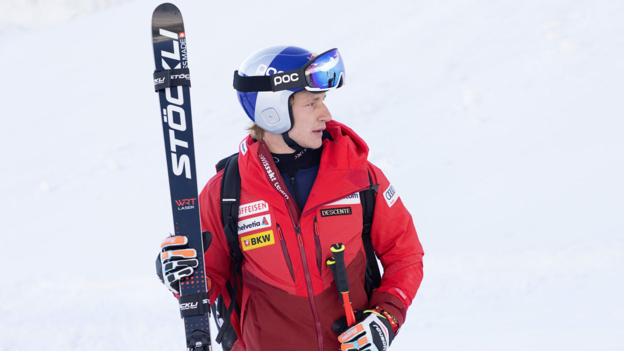 Ein Skirennfahrer in roter Team-Ausrüstung mit Helm und Skibrille steht im Schnee und hält einen schwarzen Stöckli-Ski sowie Skistöcke in den Händen.